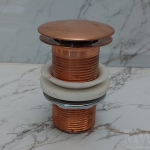 Puede incluir: Un tapón de desagüe de lavabo de baño de color cobre con un anillo de plástico blanco alrededor de la base. El tapón es de metal y tiene un acabado liso y pulido.