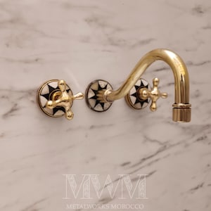 Può includere: Un rubinetto da parete dorato con maniglie a forma di croce e un beccuccio curvo. La base del rubinetto presenta un design a stella in bianco e nero. Il testo "METALWORKS MOROCCO" è visibile in basso.