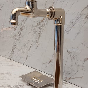 K&ouml;nnte beinhalten: Ein polierter Chrom-Wasserhahn mit einem kreuzf&ouml;rmigen Griff und einer hohen, zylindrischen Basis. Der Wasserhahn ist auf einer wei&szlig;en Marmor-Arbeitsplatte mit einem goldfarbenen Waschbecken montiert.