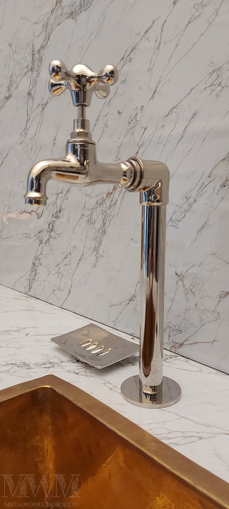 K&ouml;nnte beinhalten: Ein polierter Chrom-Wasserhahn mit einem kreuzf&ouml;rmigen Griff und einer hohen, zylindrischen Basis. Der Wasserhahn ist auf einer wei&szlig;en Marmor-Arbeitsplatte mit einem goldfarbenen Waschbecken montiert.