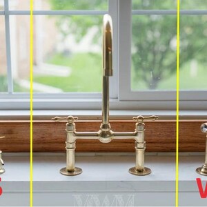 May include: A gold kitchen faucet with two handles and a sprayer. The faucet is mounted on a white countertop with a brown wooden backsplash. The faucet is labeled "W" and there is a soap dispenser labeled "S".