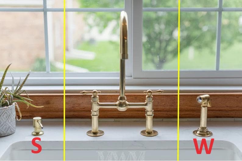 May include: A gold kitchen faucet with two handles and a sprayer. The faucet is mounted on a white countertop with a brown wooden backsplash. The faucet is labeled "W" and there is a soap dispenser labeled "S".