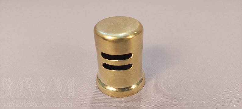 May include: A gold-colored metal vent cover with two horizontal slots. The vent cover is cylindrical and has a smooth, polished finish.