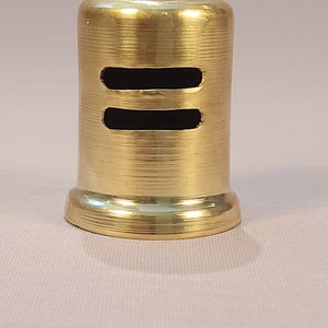 May include: A gold metal bottle stopper with two horizontal slits. The stopper has a smooth, shiny finish.