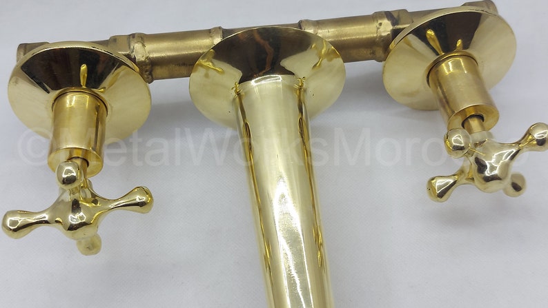 May include: A gold-toned faucet with two cross-shaped handles and a central spout. The faucet is wall-mounted and has a polished finish.