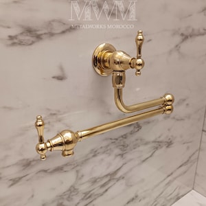 Może przedstawiać: Złoty, montowany na ścianie element wyposażenia łazienki o dekoracyjnym wzornictwie. Element wyposażenia obejmuje kran i podwójny wieszak na ręczniki. Tekst "MWM METALWORKS MOROCCO" jest widoczny u góry obrazu.