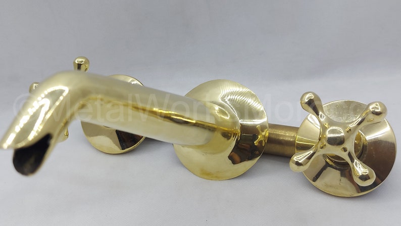 May include: A gold-toned brass faucet with two cross-shaped handles. The faucet has a curved spout and is designed for wall mounting.