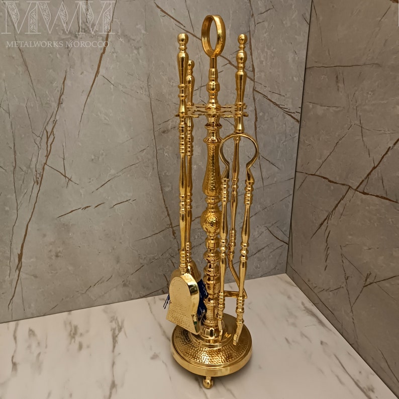 May include: A gold metal fireplace tool set with a stand. The set includes a poker, shovel, brush, and tongs. The stand has a hammered finish.