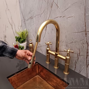 May include: A gold-colored kitchen faucet with a pull-down sprayer. The faucet has a bridge design with cross-shaped handles. The sink is copper-colored, and the background features a marble-patterned wall.