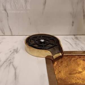 K&ouml;nnte beinhalten: Ein goldfarbenes Metallwaschbecken mit einem schwarzen Metallrost und einem Auslauf. Das Waschbecken ist in eine wei&szlig;e Marmorarbeitsplatte eingelassen.