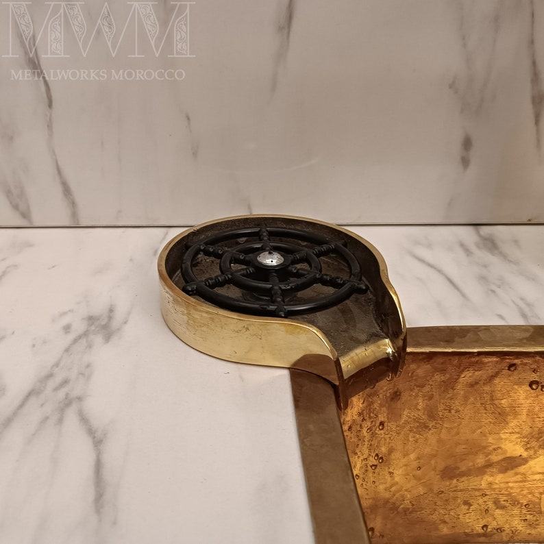 K&ouml;nnte beinhalten: Ein goldfarbenes Metallwaschbecken mit einem schwarzen Metallrost und einem Auslauf. Das Waschbecken ist in eine wei&szlig;e Marmorarbeitsplatte eingelassen.