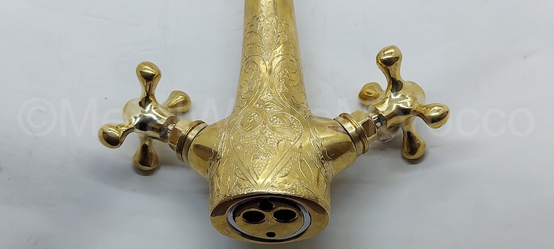 Op de afbeelding: Een goudkleurige kraan met een sierlijk bloemmotief. De kraan heeft twee kruisvormige handvatten en een tuit met drie gaten. Het ontwerp is gedetailleerd en elegant, geschikt voor een badkamer of keuken.