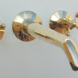 May include: A gold-toned wall-mounted faucet with two cross-shaped handles. The faucet has a curved spout and a polished finish.