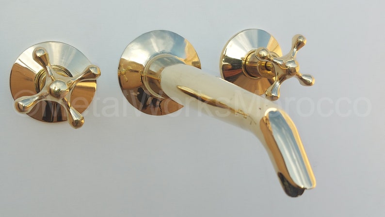 May include: A gold-toned wall-mounted faucet with two cross-shaped handles. The faucet has a curved spout and a polished finish.