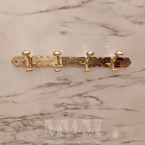 May include: A brass wall-mounted coat rack with four hooks. The rack has a decorative, textured design and is mounted on a marble-patterned wall. The hooks are evenly spaced and have a rounded, knob-like shape. The text "METALWORKS MOROCCO" is at the bottom.