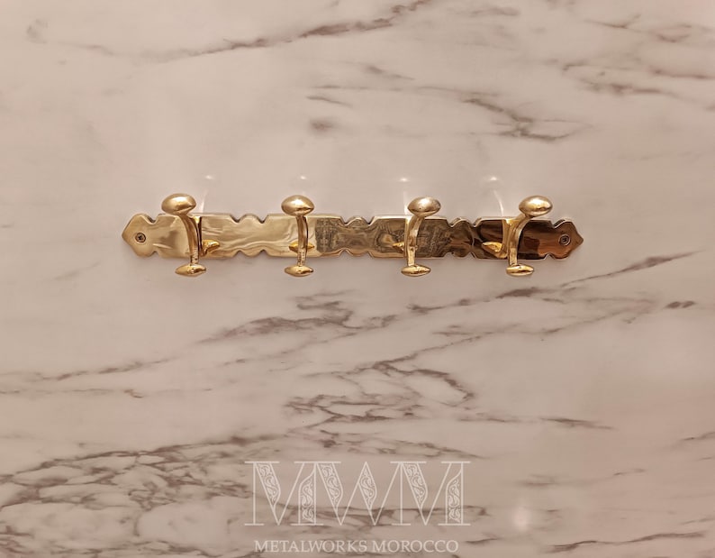 May include: A brass wall-mounted coat rack with four hooks. The rack has a decorative, textured design and is mounted on a marble-patterned wall. The hooks are evenly spaced and have a rounded, knob-like shape. The text "METALWORKS MOROCCO" is at the bottom.
