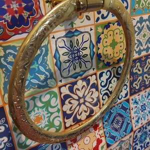 K&ouml;nnte beinhalten: Ein goldfarbener Handtuchring mit einem verzierten Design, montiert an einer wei&szlig;en gekachelten Wand mit bunten Mustern.