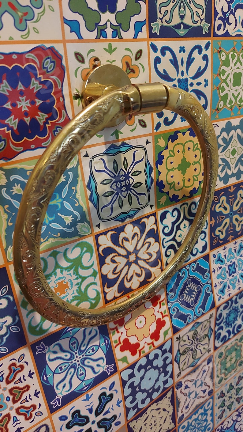 K&ouml;nnte beinhalten: Ein goldfarbener Handtuchring mit einem verzierten Design, montiert an einer wei&szlig;en gekachelten Wand mit bunten Mustern.