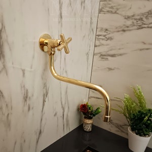 May include: A gold-toned wall-mounted faucet with a cross-shaped handle. The faucet has a long, curved spout and is mounted on a white marble wall.