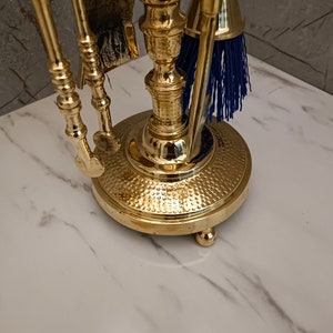 May include: A gold metal fireplace tool set with a brush, shovel, and poker. The tools are mounted on a decorative stand with a hammered finish.
