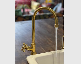 May include: A gold-toned faucet with a cross-shaped handle is mounted on a wooden countertop. Water is flowing from the spout into a white porcelain sink.