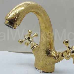 Op de afbeelding: Een goudkleurige kraan met een sierlijk bloemmotief. De kraan heeft een gebogen uitloop en twee kruisvormige handgrepen. De afbeelding toont de kraan tegen een witte achtergrond. De tekst "MetalWork Morocco" is zichtbaar.