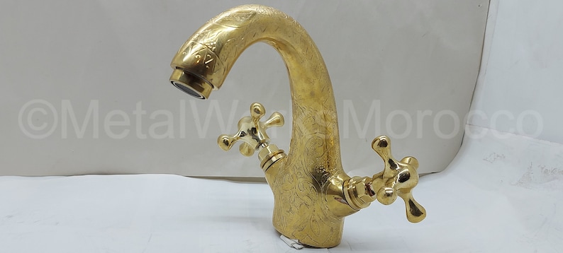 Op de afbeelding: Een goudkleurige kraan met een sierlijk bloemmotief. De kraan heeft een gebogen uitloop en twee kruisvormige handgrepen. De afbeelding toont de kraan tegen een witte achtergrond. De tekst "MetalWork Morocco" is zichtbaar.