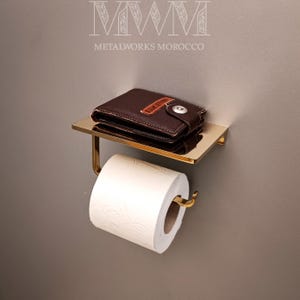 Może przedstawiać: Złoty metalowy uchwyt na papier toaletowy z małą półką. Na półce leży brązowy skórzany portfel. Rolka białego papieru toaletowego jest na miejscu. U góry znajduje się napis "METALWORKS MOROCCO".