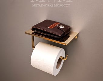 May include: A gold-coloured metal toilet roll holder with a small shelf. A brown leather wallet rests on the shelf. A roll of white toilet paper is in place. The text "METALWORKS MOROCCO" is at the top.