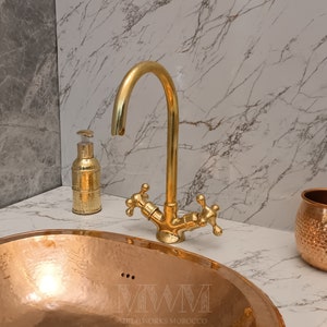 May include: A gold-toned faucet with two handles is mounted on a white marble countertop. A copper bowl sink is in the foreground. The faucet is labeled "MWM Metalworks Morocco".