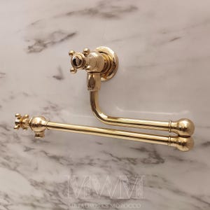 Könnte beinhalten: Ein goldfarbener, an der Wand montierter Handtuchhalter mit kreuzförmigem Griff und gebogenem Arm. Der Halter hat zwei parallele Stangen zum Aufhängen von Handtüchern. Die Armatur ist aus Metall und an einer marmorierten Wand befestigt.