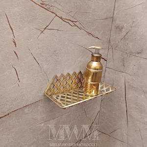 May include: Gold metal bathroom shelf with a geometric design. A gold bottle of soap is on the shelf. The shelf is mounted on a light gray tiled wall.