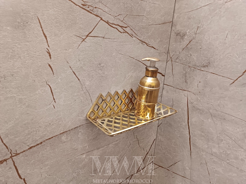 May include: Gold metal bathroom shelf with a geometric design. A gold bottle of soap is on the shelf. The shelf is mounted on a light gray tiled wall.
