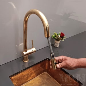 May include: A gold kitchen faucet with a pull-down sprayer. The faucet is installed on a copper sink. The sprayer is being pulled out by a hand.