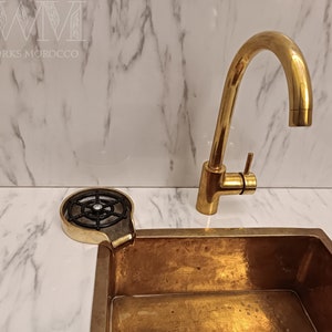 K&ouml;nnte beinhalten: Ein goldfarbenes K&uuml;chensp&uuml;lbecken mit passendem Wasserhahn und einem schwarzen Abflusssieb aus Metall. Das Sp&uuml;lbecken ist auf einer wei&szlig;en Marmor-Arbeitsplatte montiert.