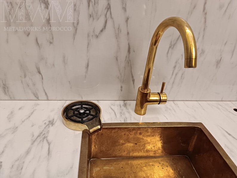 K&ouml;nnte beinhalten: Ein goldfarbenes K&uuml;chensp&uuml;lbecken mit passendem Wasserhahn und einem schwarzen Abflusssieb aus Metall. Das Sp&uuml;lbecken ist auf einer wei&szlig;en Marmor-Arbeitsplatte montiert.