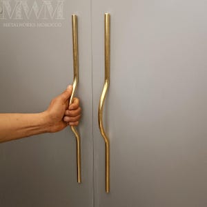 May include: Two gold-toned cabinet door handles with a curved design. The handles are mounted on a grey wall.