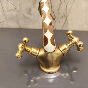 May include: A brass faucet featuring a curved spout with an inlaid pattern of white and brown wood diamond shapes. The faucet has two cross-shaped handles and a polished gold-tone finish. The text METALWORKS MOROCCO is visible in the image.
