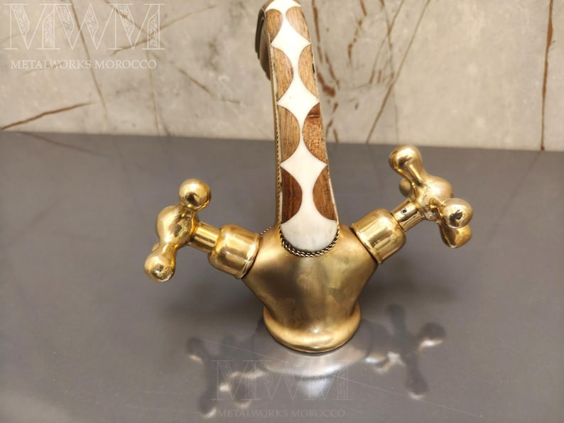 May include: A brass faucet featuring a curved spout with an inlaid pattern of white and brown wood diamond shapes. The faucet has two cross-shaped handles and a polished gold-tone finish. The text METALWORKS MOROCCO is visible in the image.