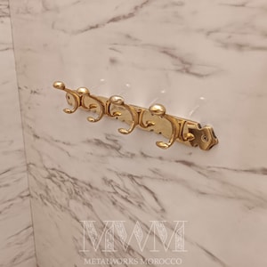 May include: A gold-colored metal wall-mounted coat rack with five curved hooks. The rack is mounted on a marble-patterned wall. The text "METALWORKS MOROCCO" is at the bottom.