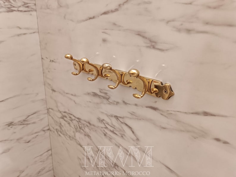 May include: A gold-colored metal wall-mounted coat rack with five curved hooks. The rack is mounted on a marble-patterned wall. The text "METALWORKS MOROCCO" is at the bottom.