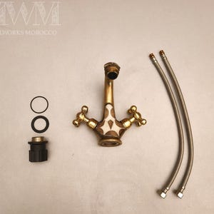 May include: A brass bathroom faucet with two cross-shaped handles and a central white inlay featuring a geometric pattern. The set includes two flexible metal supply hoses, two black rubber O-rings, and a black plastic mounting component.