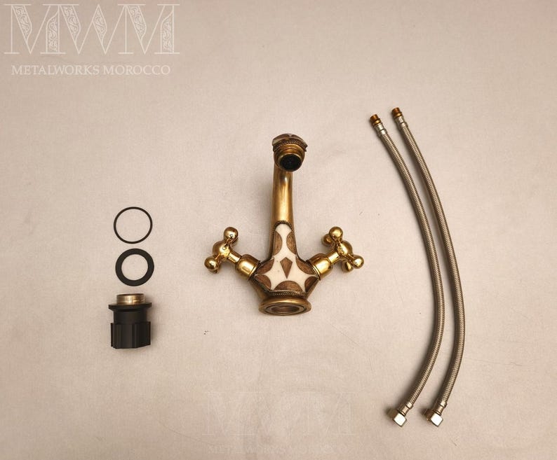 May include: A brass bathroom faucet with two cross-shaped handles and a central white inlay featuring a geometric pattern. The set includes two flexible metal supply hoses, two black rubber O-rings, and a black plastic mounting component.