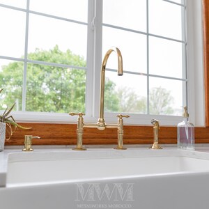May include: A gold kitchen faucet with a high arc spout and two handles. The faucet is installed on a white countertop in front of a window with a wooden frame.