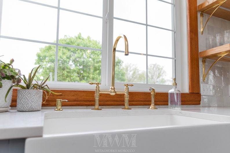 May include: A gold kitchen faucet with a high arc spout and two handles. The faucet is installed on a white countertop in front of a window with a wooden frame.