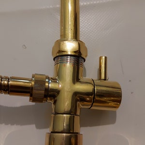 Puede incluir: Primer plano de un grifo dorado con una manguera y una v&aacute;lvula desviadora. El grifo tiene un acabado pulido y un dise&ntilde;o sencillo.