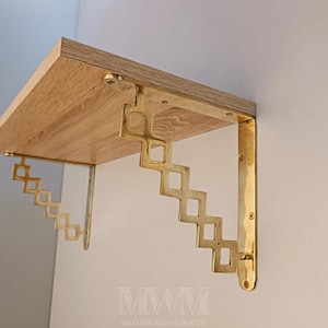 Puede incluir: Una estantería de madera con soportes metálicos de color latón. Los soportes tienen un diseño geométrico escalonado. La estantería está montada en una pared de color claro. Los soportes metálicos están fijados a la estantería con tornillos.