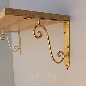 Op de afbeelding: Twee gouden metalen plankbeugels met sierlijke krulontwerpen. De beugels zijn bevestigd aan een houten plank.