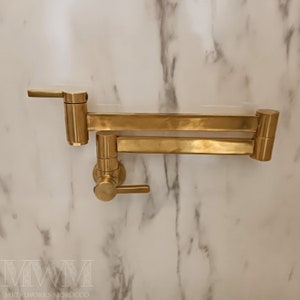 May include: A gold-toned kitchen faucet with a double-jointed arm, mounted on a white marble wall. The faucet has two handles, one on each side of the arm.