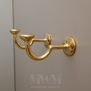 Könnte beinhalten: Zwei goldfarbene Metallhaken, die an einer Wand montiert sind. Die Haken sind gebogen und haben eine abgerundete Oberseite. Die Haken sind mit Schrauben an der Wand befestigt.